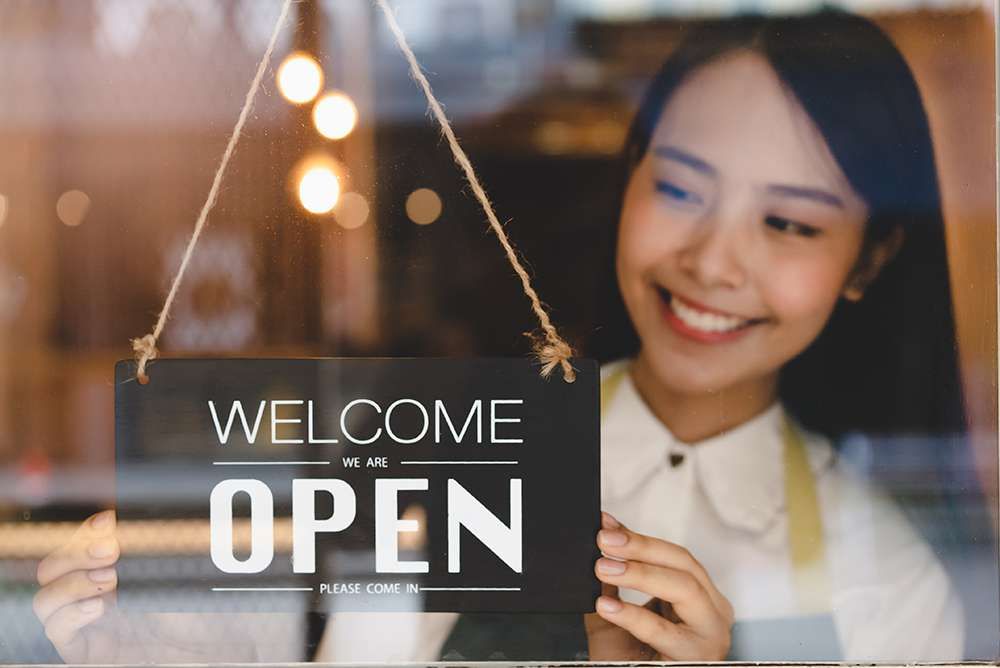 A smiling shop worker displays a hanging sign reading WELCOME WE ARE OPEN.
