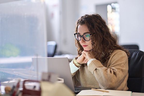 A woman in glasses looks at a laptop with her hands clasped beneath her chin.