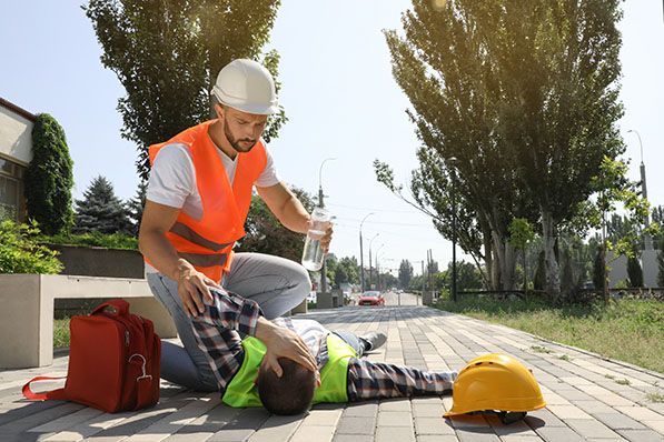 A worker kneels beside a coworker lying on pavement, with a hard hat and first-aid bag nearby.