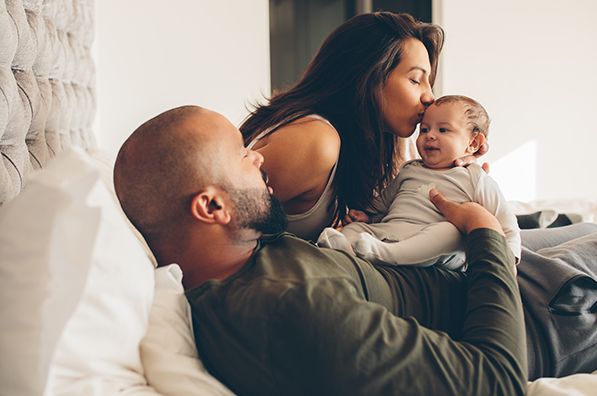 Two adults cuddle a baby on a bed, with one kissing the baby's head.