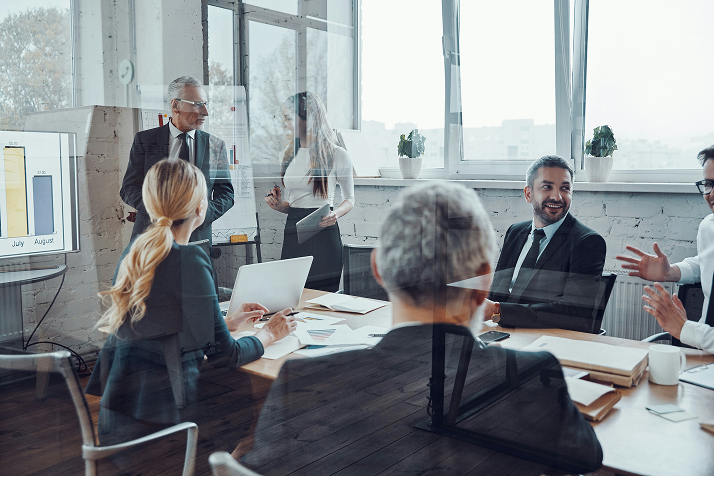 Businesspeople meet around a table with laptops in a bright, glass-walled conference room.