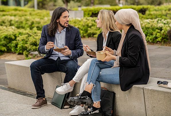 Three coworkers sit outdoors sharing conversation while eating from food containers.