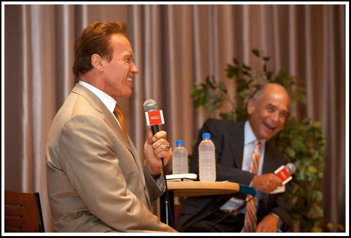 A man in a light-colored suit speaks into a microphone while another seated man laughs.