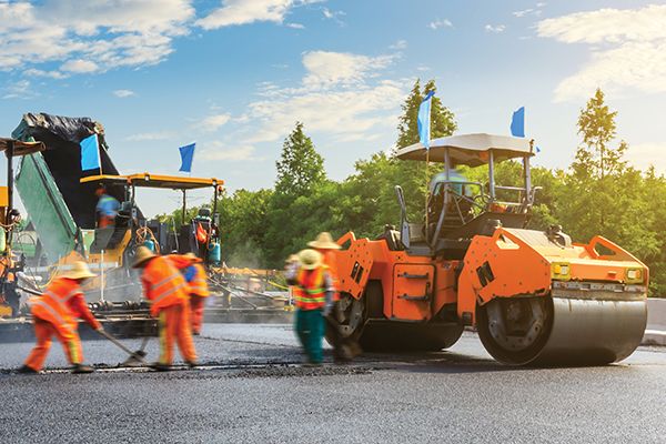 Road workers in orange safety clothing pave a road beside a large roller.