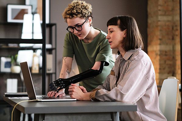 Two women are working together on a laptop, one using a prosthetic arm to type.