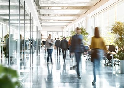 People walk through a bright office corridor lined with glass walls and plants.