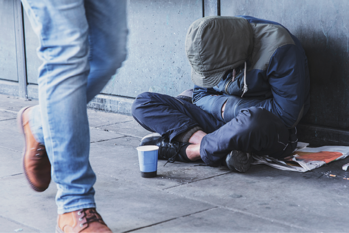 A person in a hooded jacket sits curled up on a sidewalk beside a cup as a pedestrian passes.