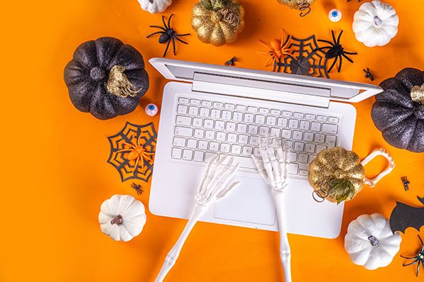 Skeleton hands typing on a laptop keyboard, surrounded by Halloween pumpkins, spiders, and webs on an orange background.