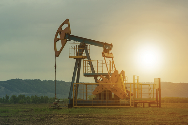 An oil pumpjack stands in a field with the low sun behind it.