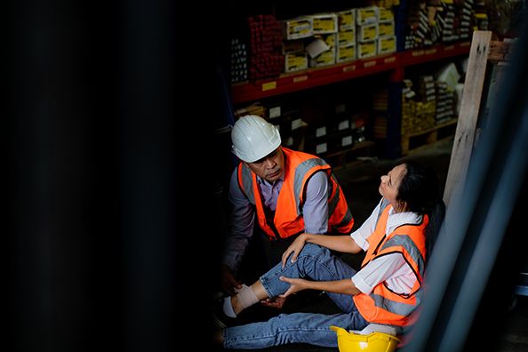 A worker in a hard hat kneels beside a coworker seated on a warehouse floor.