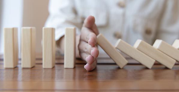 A hand stops a row of falling wooden dominoes from reaching the upright pieces.
