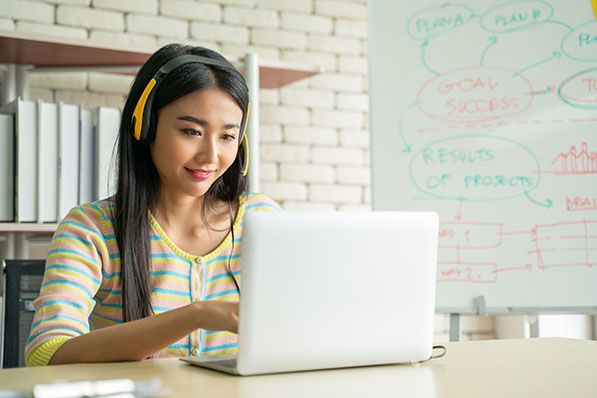 A woman wearing headphones works on a laptop beside a whiteboard.