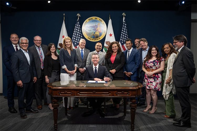 A group stands around a man seated at a signing desk beneath an official seal and flags.