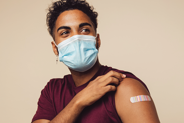 A masked person pulls up a sleeve to show a small adhesive bandage on the upper arm.