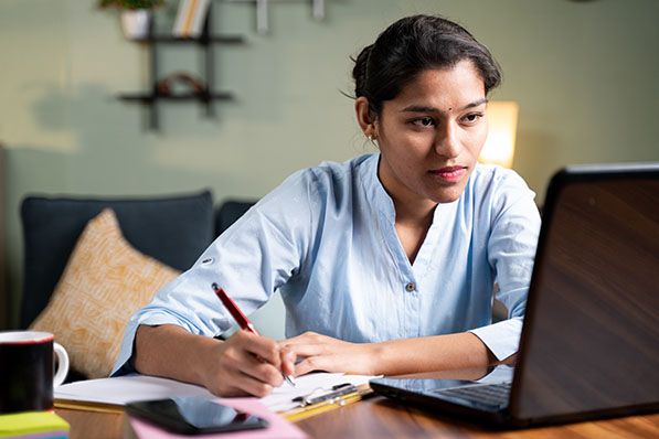 A woman writes in a notebook while looking at a laptop at a desk.