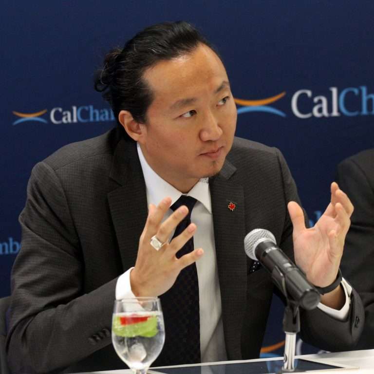 Brandon A. Lee gestures while seated at a table with a microphone before a CalChamber backdrop.