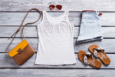 A sleeveless top, denim shorts, sandals, sunglasses and a handbag arranged on wooden boards.