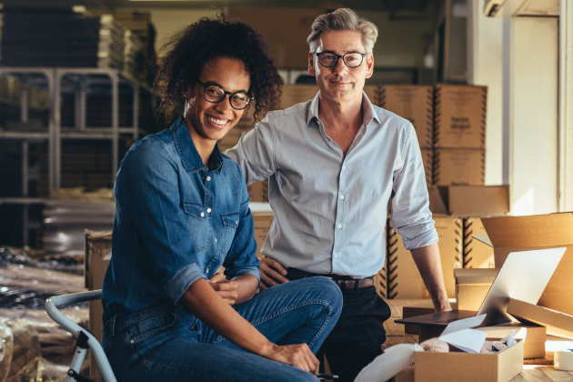 Two people smile beside a laptop and packing boxes in a workshop.