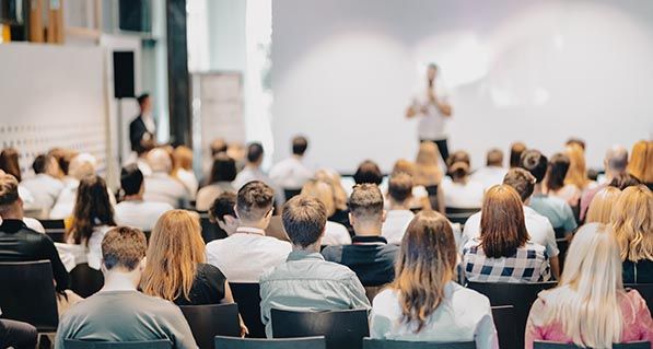 Audience members face a speaker at the front of a conference room.