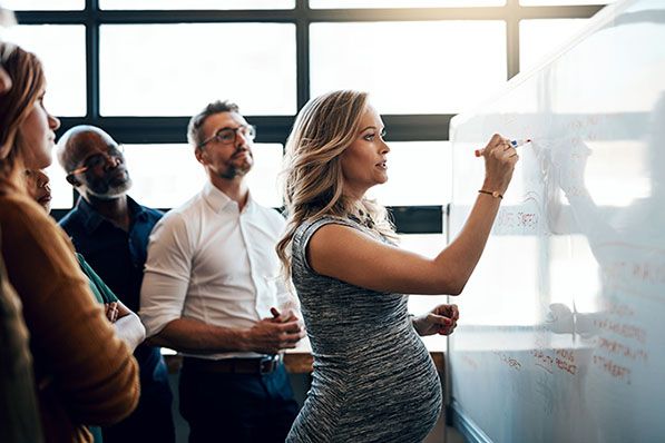 A pregnant woman writes on a whiteboard while colleagues watch.