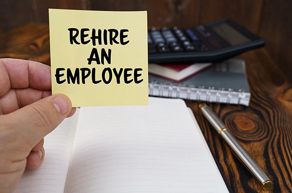 A hand holds a yellow note reading REHIRE AN EMPLOYEE above a notebook and calculator.