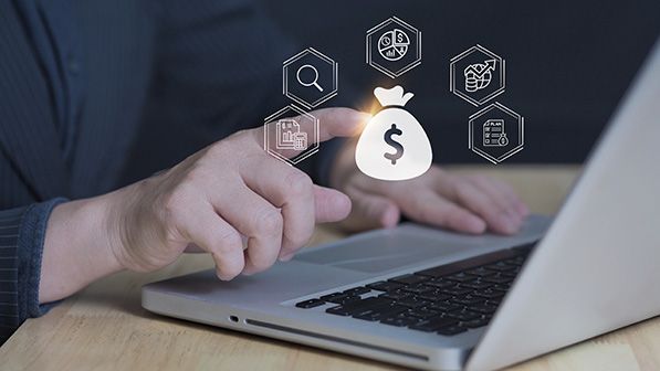 A person uses a laptop beneath overlaid financial icons surrounding a dollar-sign money bag.