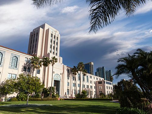 A tall civic building and palm trees stand beneath a dramatic cloudy sky.