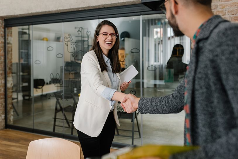 A smiling woman in a white blazer shakes hands with a person in an office.