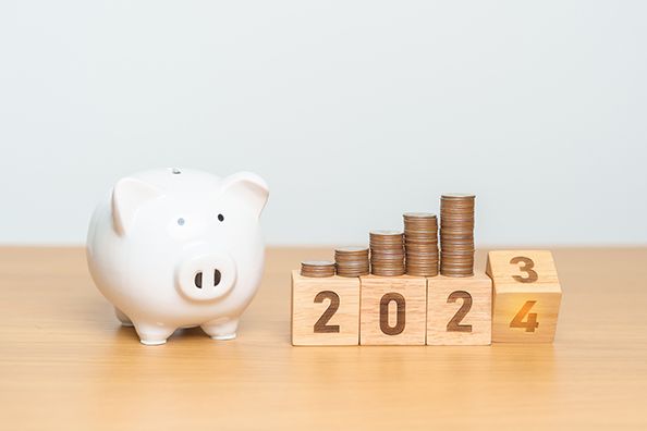 A white piggy bank beside coin stacks on wooden calendar blocks changing from 2023 to 2024.