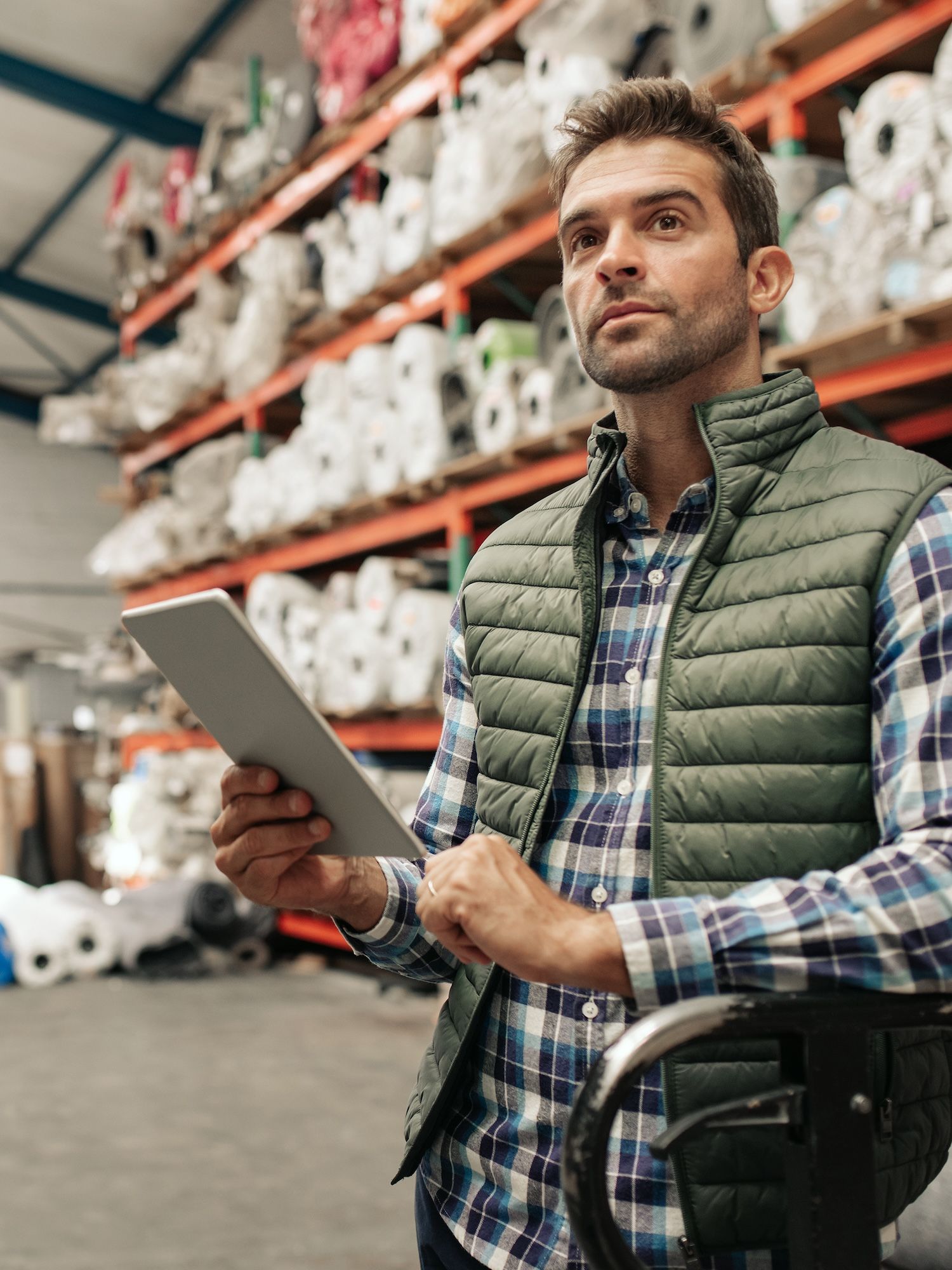 A warehouse worker holds a tablet beside shelves stacked with rolls of material.