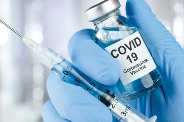 A gloved hand holds a vial labeled COVID 19 Coronavirus Vaccine beside a syringe.