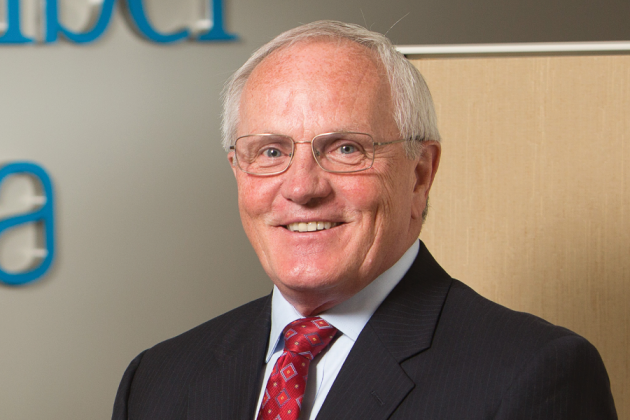 Portrait of a smiling white-haired man wearing glasses, a dark suit and red patterned tie.