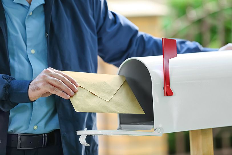 A person places an envelope into a white roadside mailbox with a red flag.