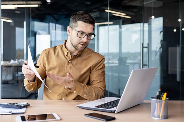 A man holds a document and gestures toward an open laptop in an office.