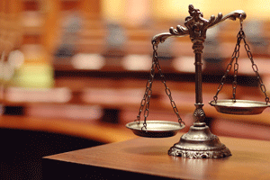 An ornate balance scale stands on a desk with blurred law books behind it.