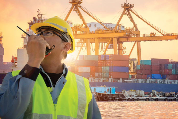 A port worker wearing a hard hat and reflective vest uses a radio near container cranes.