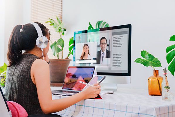 A woman wearing headphones joins a video call at a home desk with a monitor and laptop.
