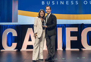 Two people pose with an award on a stage with large letters and a Business Outlook backdrop.