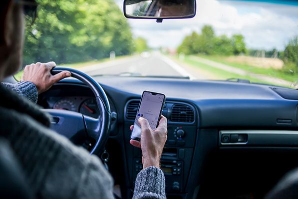 A driver holds a smartphone in one hand while steering with the other.