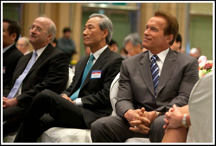 Three men in suits sit in the audience at an indoor event.