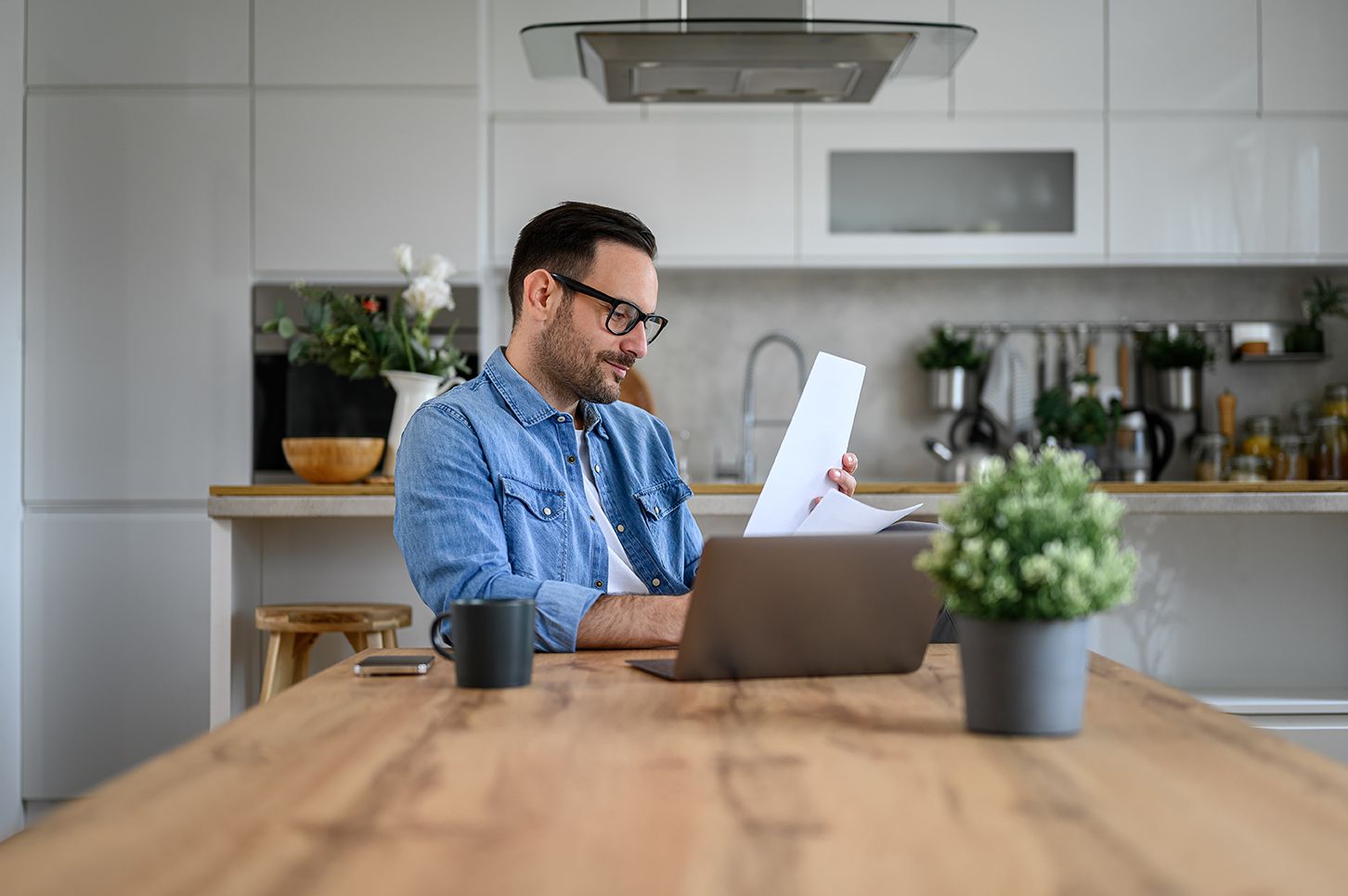 A man wearing glasses reviews papers beside a laptop at a kitchen table.