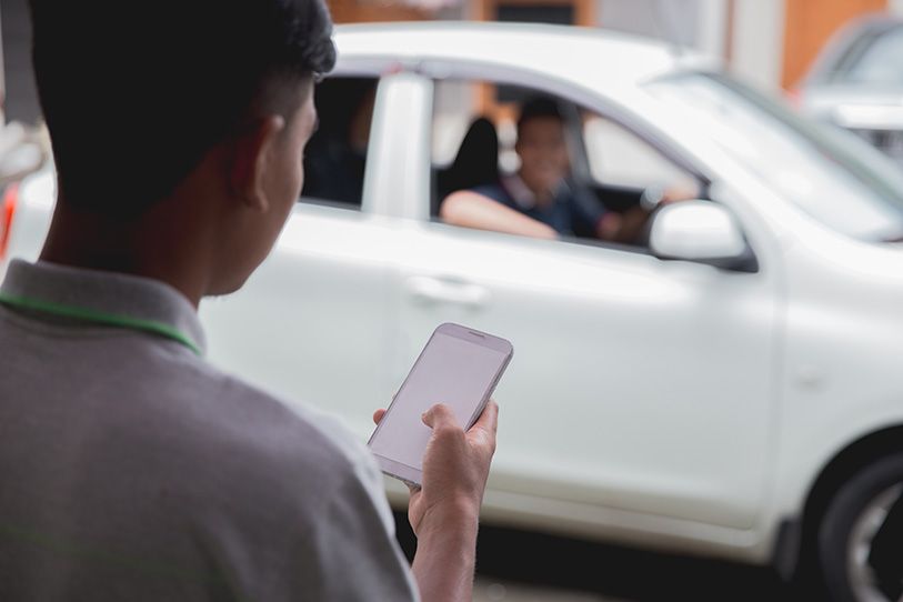 A person looks at a smartphone near a white car with its driver visible through the window.