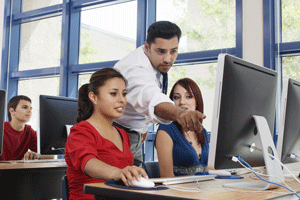 An instructor points toward a desktop monitor while students work in a computer classroom.