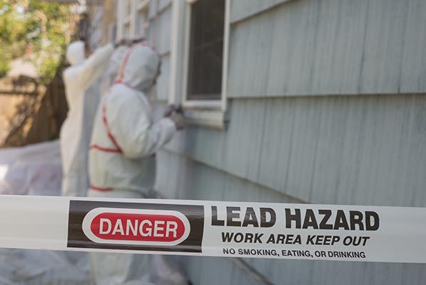 A worker in protective coveralls stands behind tape reading DANGER LEAD HAZARD, WORK AREA KEEP OUT.