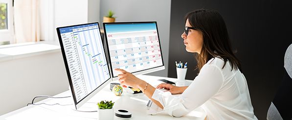 A woman works with spreadsheets and a calendar across two desktop monitors.