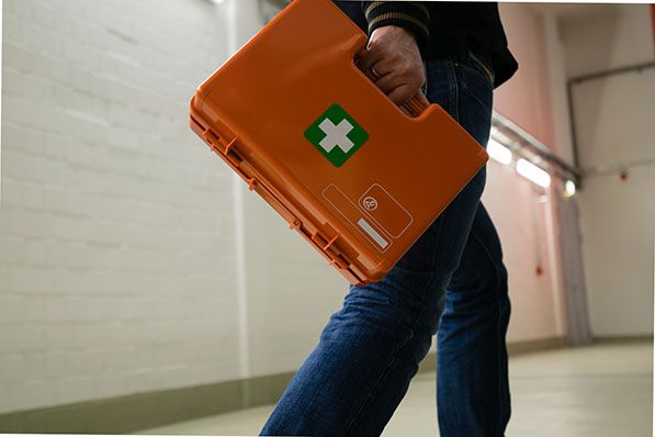 A person in jeans walks, holding an orange first aid kit with a green cross.