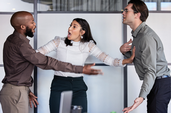 Three coworkers argue in an office, with the person in the middle extending both arms between the others.