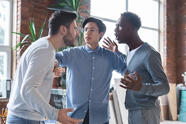 Three men stand in an office discussion, with the middle man holding his arms between the others.