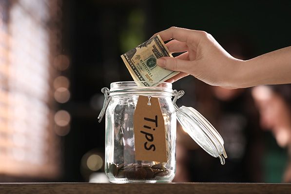 A hand drops a banknote into a glass jar labeled TIPS.