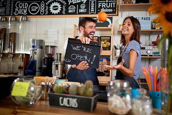 Two smiling shop workers hold a chalkboard reading WE'RE OPEN behind a counter.